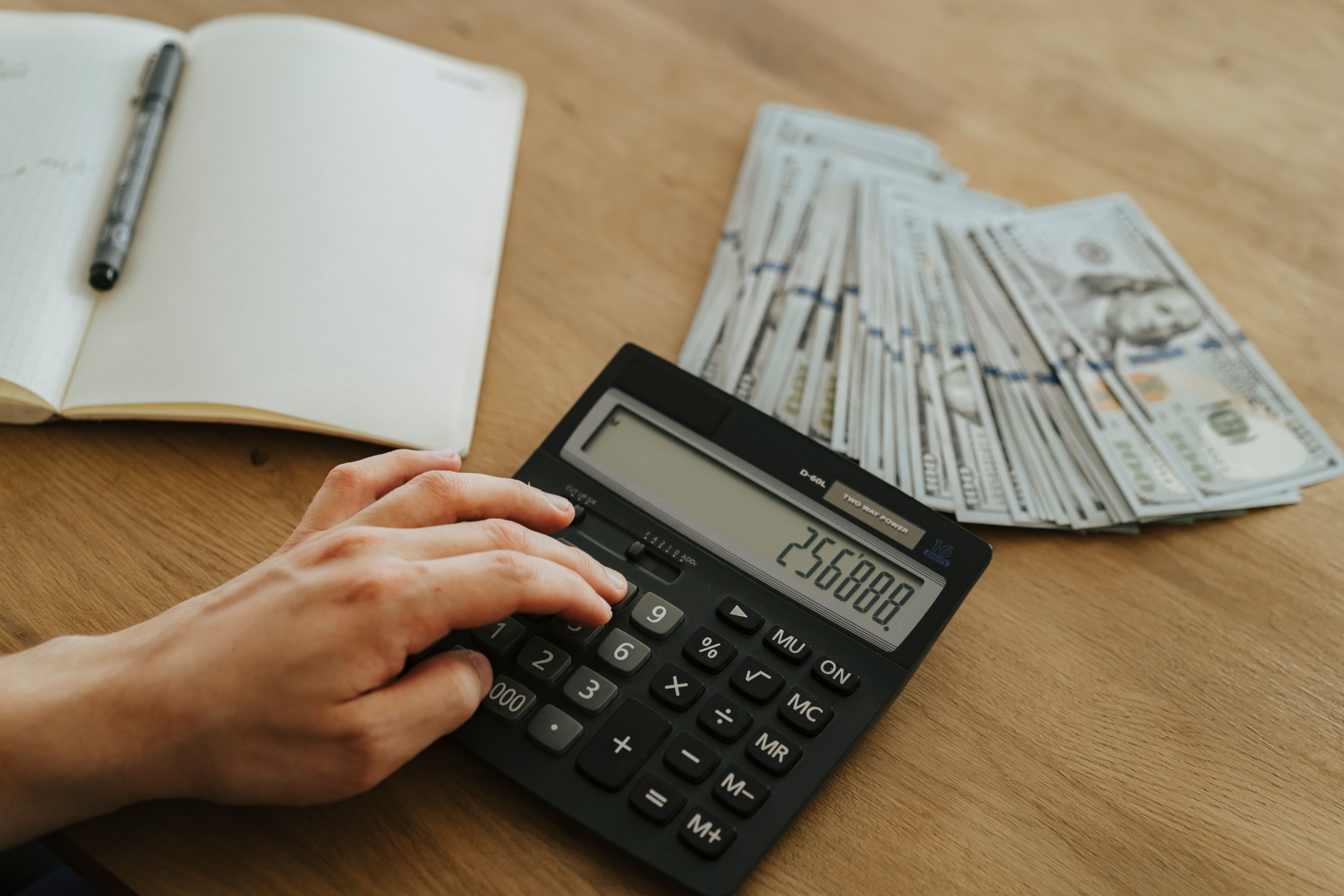 A hand using a calculator next to a notebook, pen, and a fanned stack of hundred-dollar bills on a wooden desk, representing the financial cost calculations of a workplace accident.